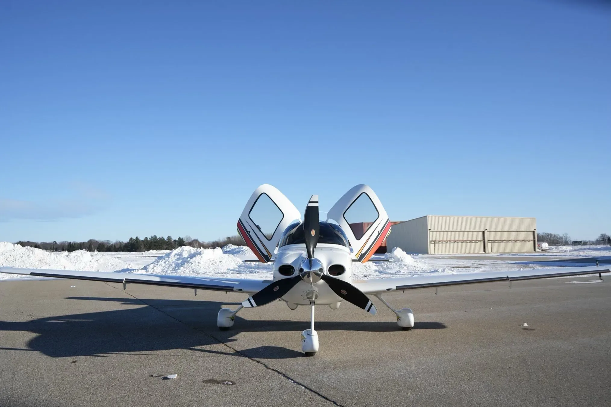 A white Cirrus SR22 aircraft with its gull-wing doors open, parked on a snowy airfield with hangars in the background.