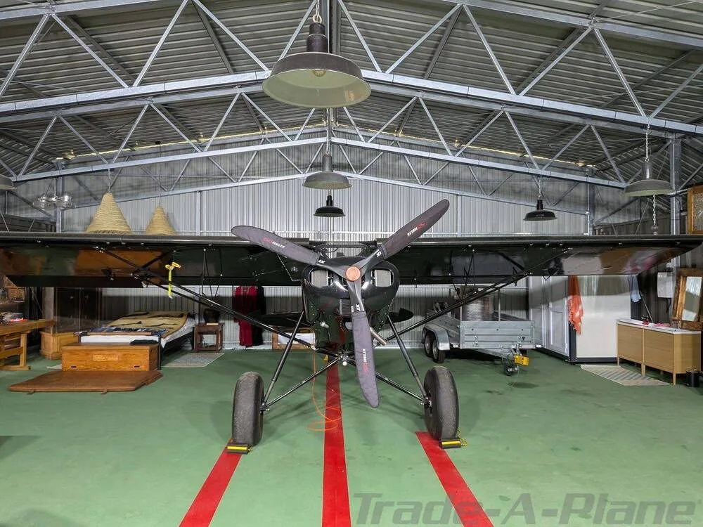 A black Zlin Shock Cub aircraft with a three-blade propeller, parked inside a hangar with a green floor.