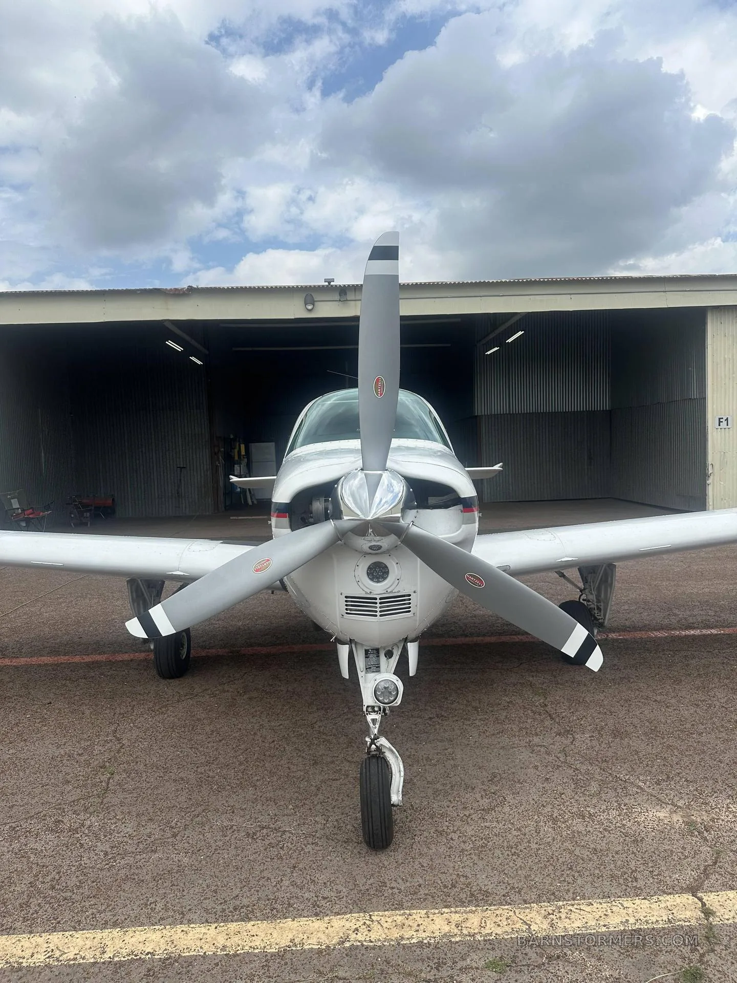 A head-on view of a white Beechcraft Debonair N6396S with a three-bladed propeller, parked in front of a hangar.