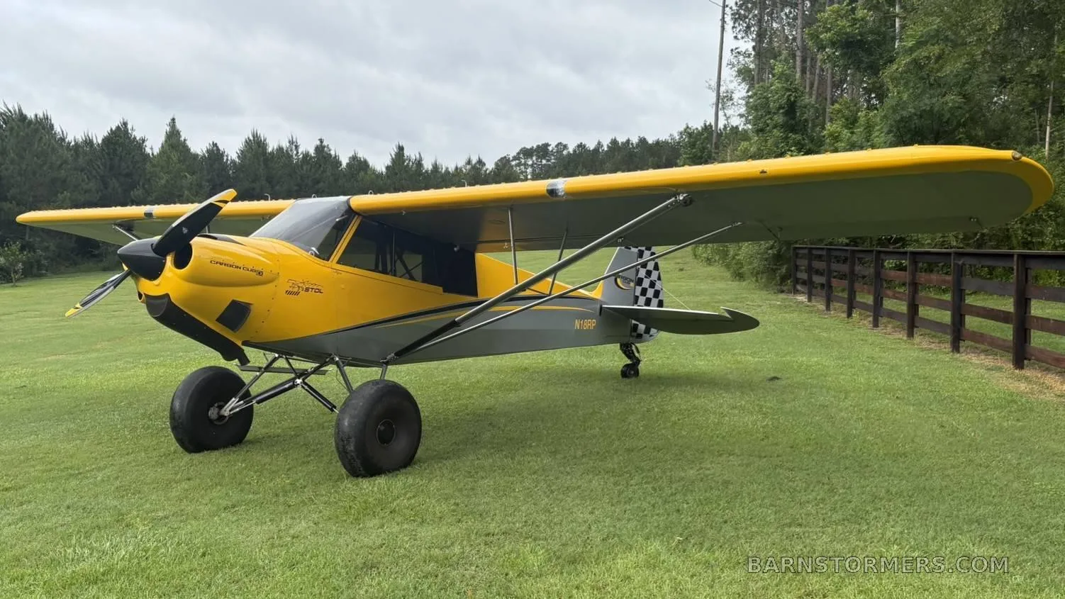 A yellow and grey CubCrafters Carbon Cub aircraft with a checkered tail, parked on a grassy field under a cloudy sky.