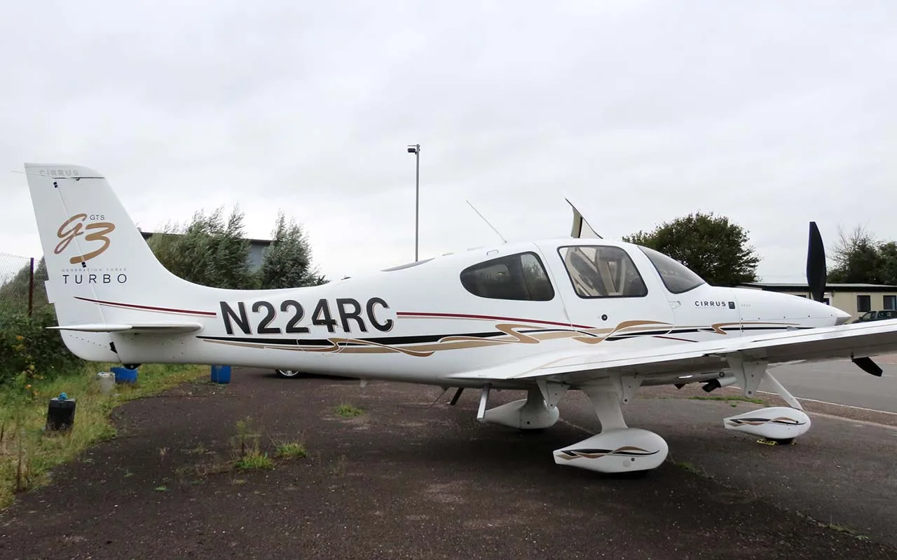 A white Cirrus SR22T (N224RC) with brown and black stripes parked on an asphalt surface next to a grassy area.