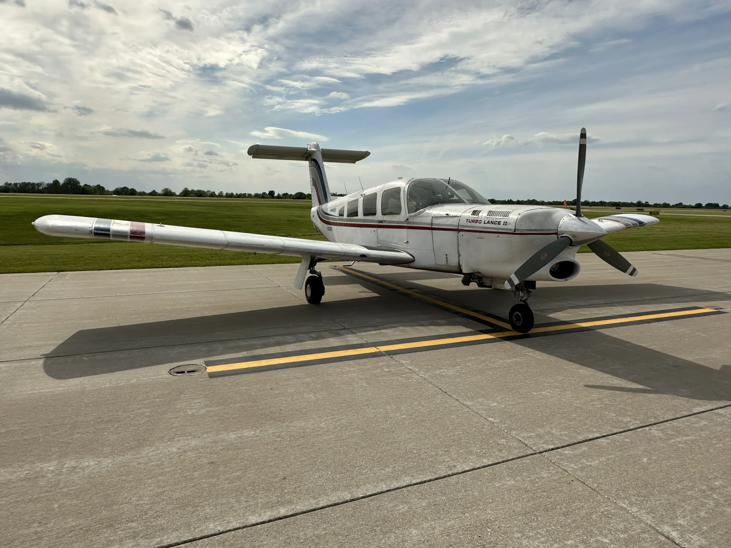 A white Piper Turbo Lance II aircraft with red and blue stripes parked on an airfield under a cloudy sky.