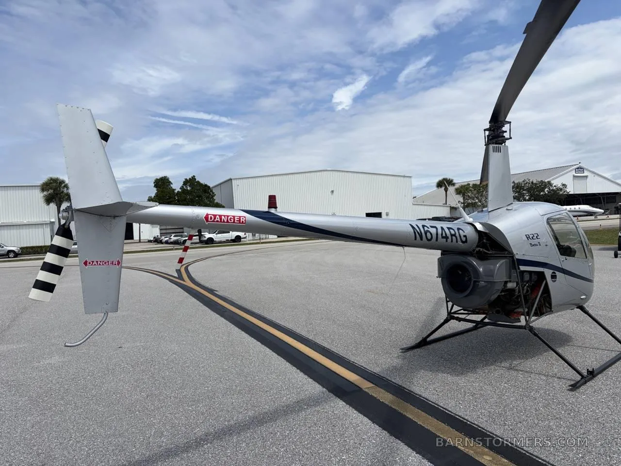 A silver Robinson R22 Beta II helicopter (N674RG) with blue stripes, viewed from the rear-right, showing the tail rotor and main rotor.