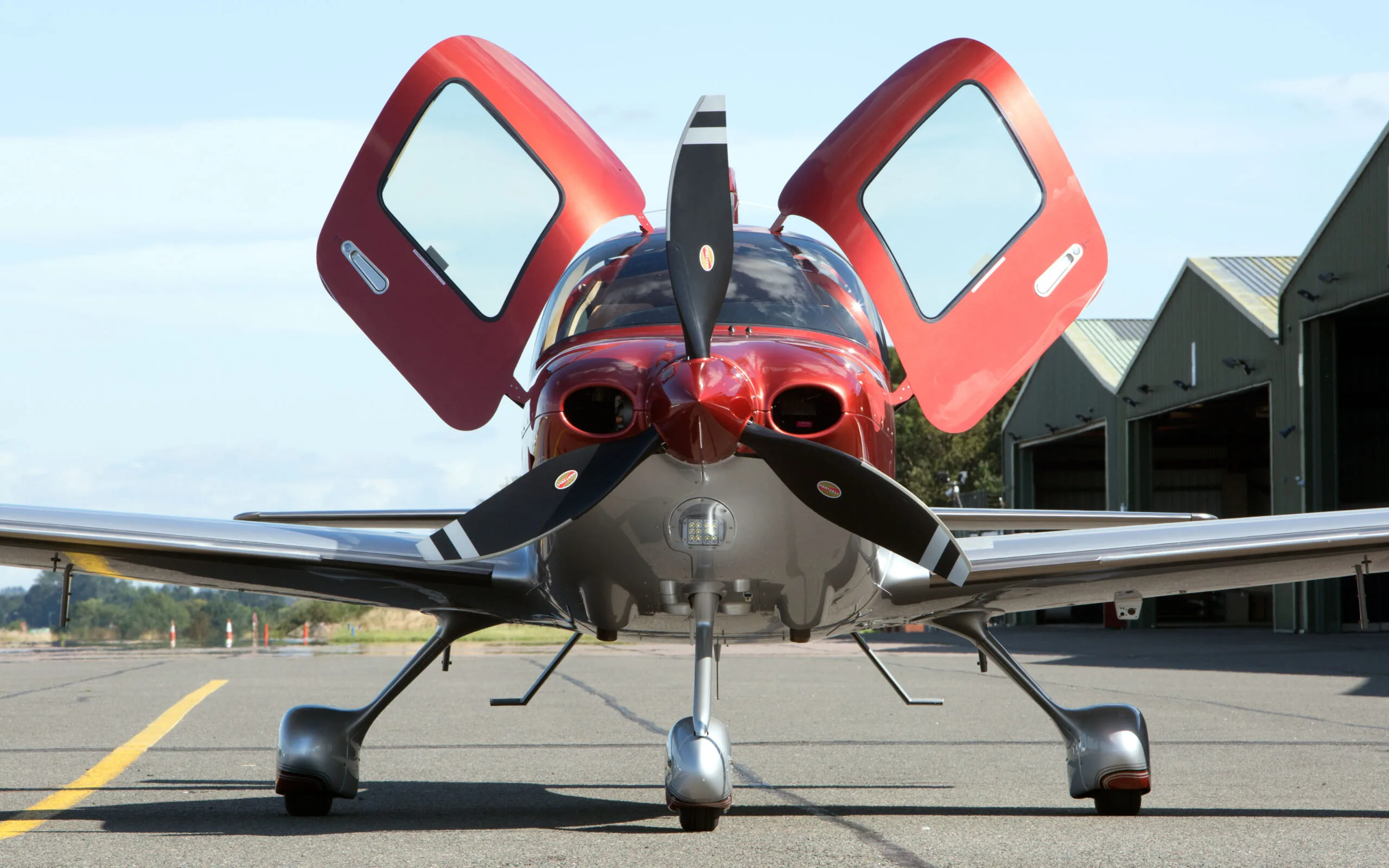 A red and silver Cirrus SR22T GTS aircraft with its gull-wing doors open, viewed head-on on an airfield tarmac with hangars behind it.