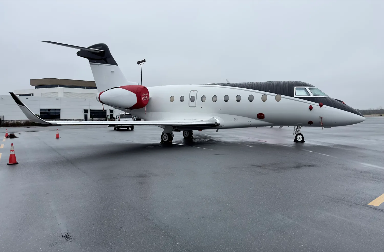 A white and black Gulfstream G280 business jet with red accents on the tail and engine nacelle, parked on a wet tarmac.