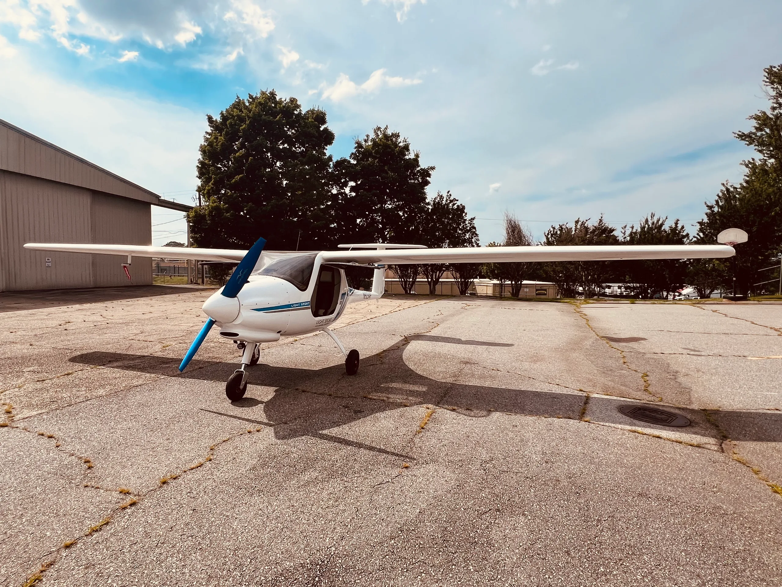 A white light sport aircraft with a blue propeller and blue stripes is parked on an asphalt apron with a hangar and trees in the background.