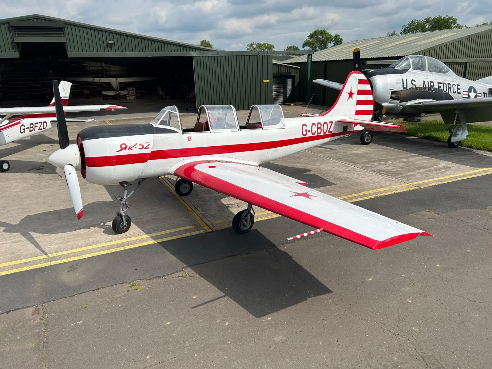 A white and red Yakovlev Yak-52 aircraft, registration G-CBOZ, is parked on an asphalt tarmac with hangars and a silver U.S. Air Force T-28 Trojan in the background.