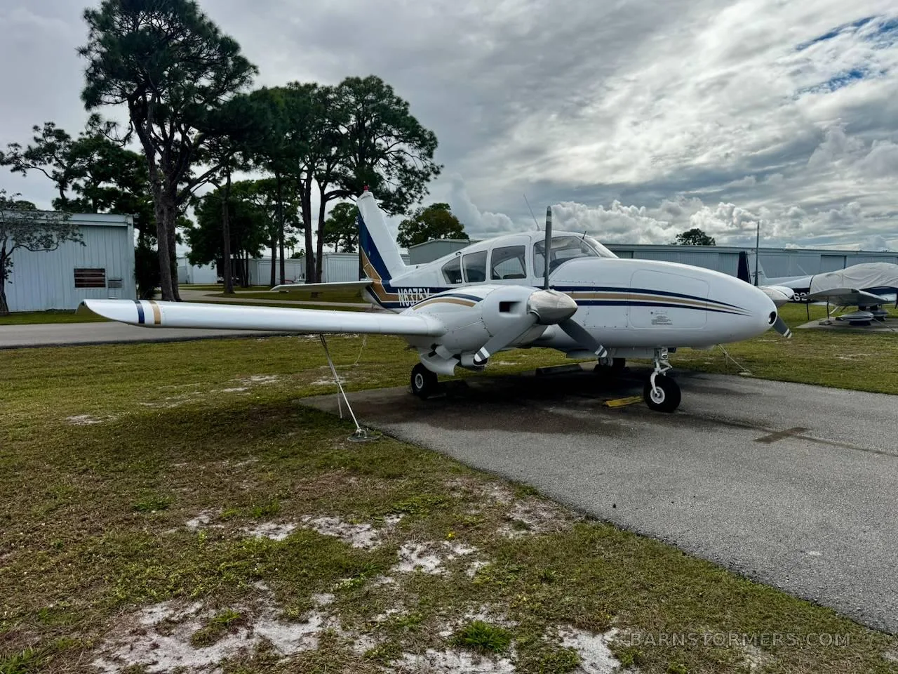 A white twin-engine aircraft with blue and gold stripes parked on an asphalt apron, with hangars and trees under a cloudy sky.