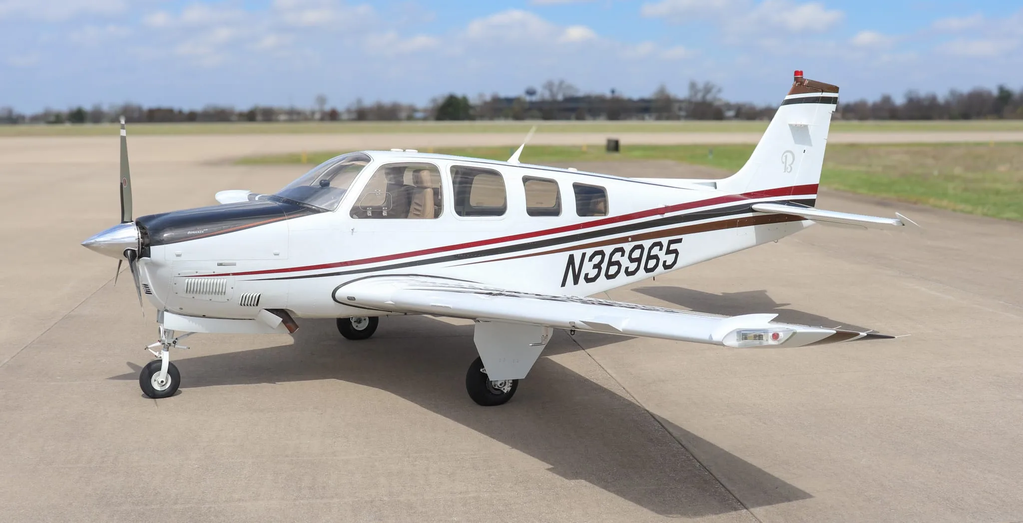 A white Beechcraft Bonanza G36 aircraft with black, red, and brown stripes, registration N36965, parked on an airfield under a partly cloudy sky.
