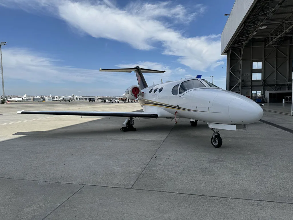 A white business jet with black and gold accents on the tail, parked on an airport apron next to a large hangar.