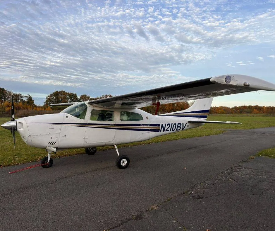 A white Cessna 210 Centurion aircraft with blue and gold stripes, registration N210BV, parked on an asphalt runway under a cloudy sky.