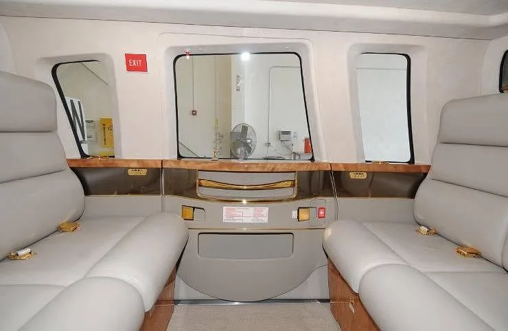 A view inside the helicopter cabin showing two rows of light gray seats facing each other, with wood and gold trim.