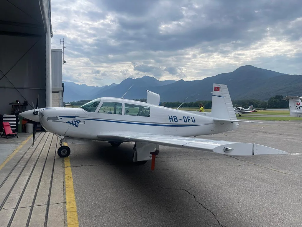 A white single-engine propeller aircraft with blue stripes and registration HB-DFU parked outside a hangar with mountains in the background.