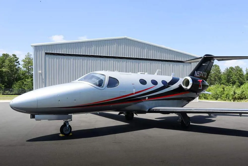A silver, black, and red Cessna Citation Mustang jet, tail number N574TA, parked on an tarmac with a hangar in the background.