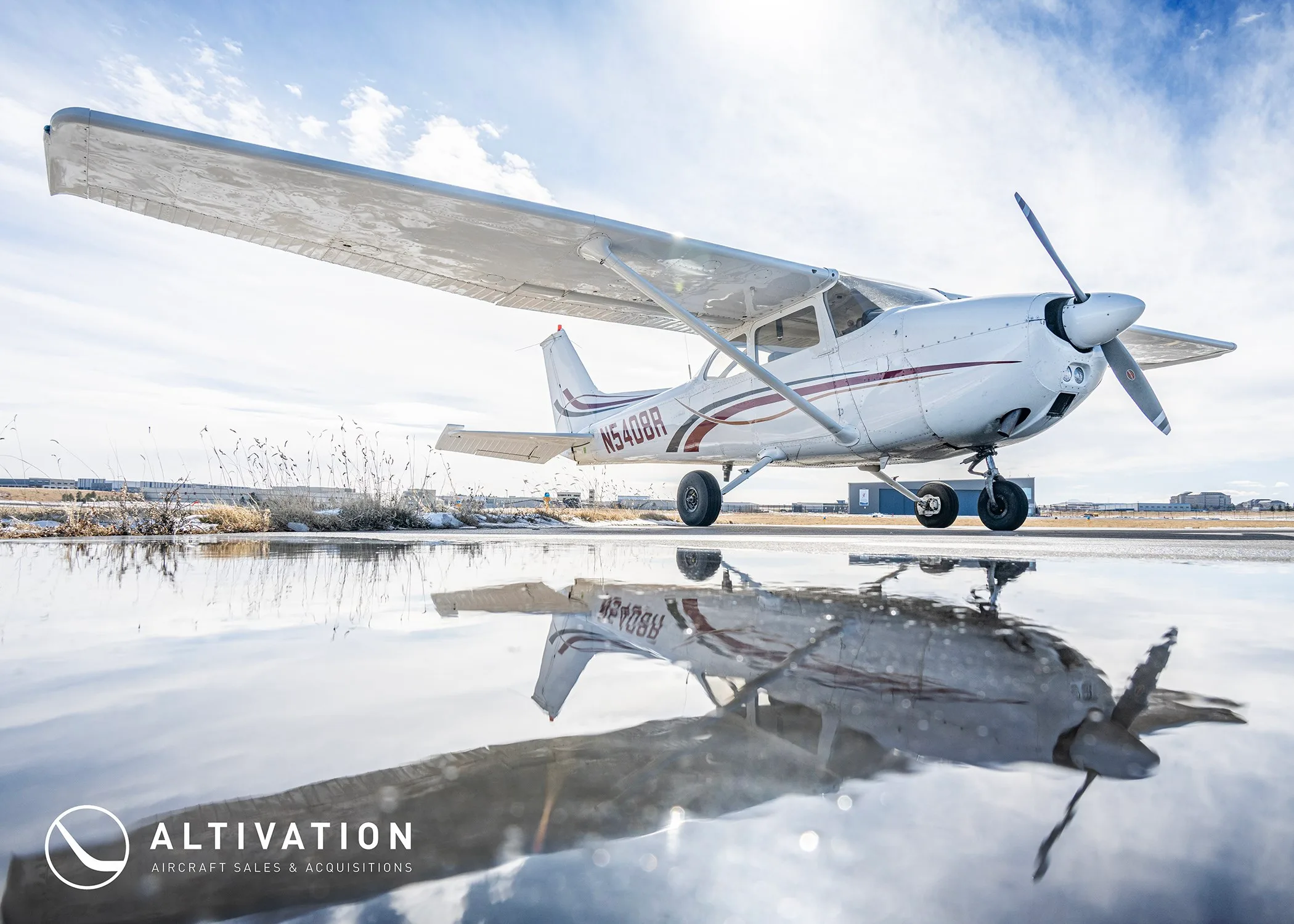 A white Cessna 172 with red and black stripes, registration N5408R, reflected in a puddle on an airfield.