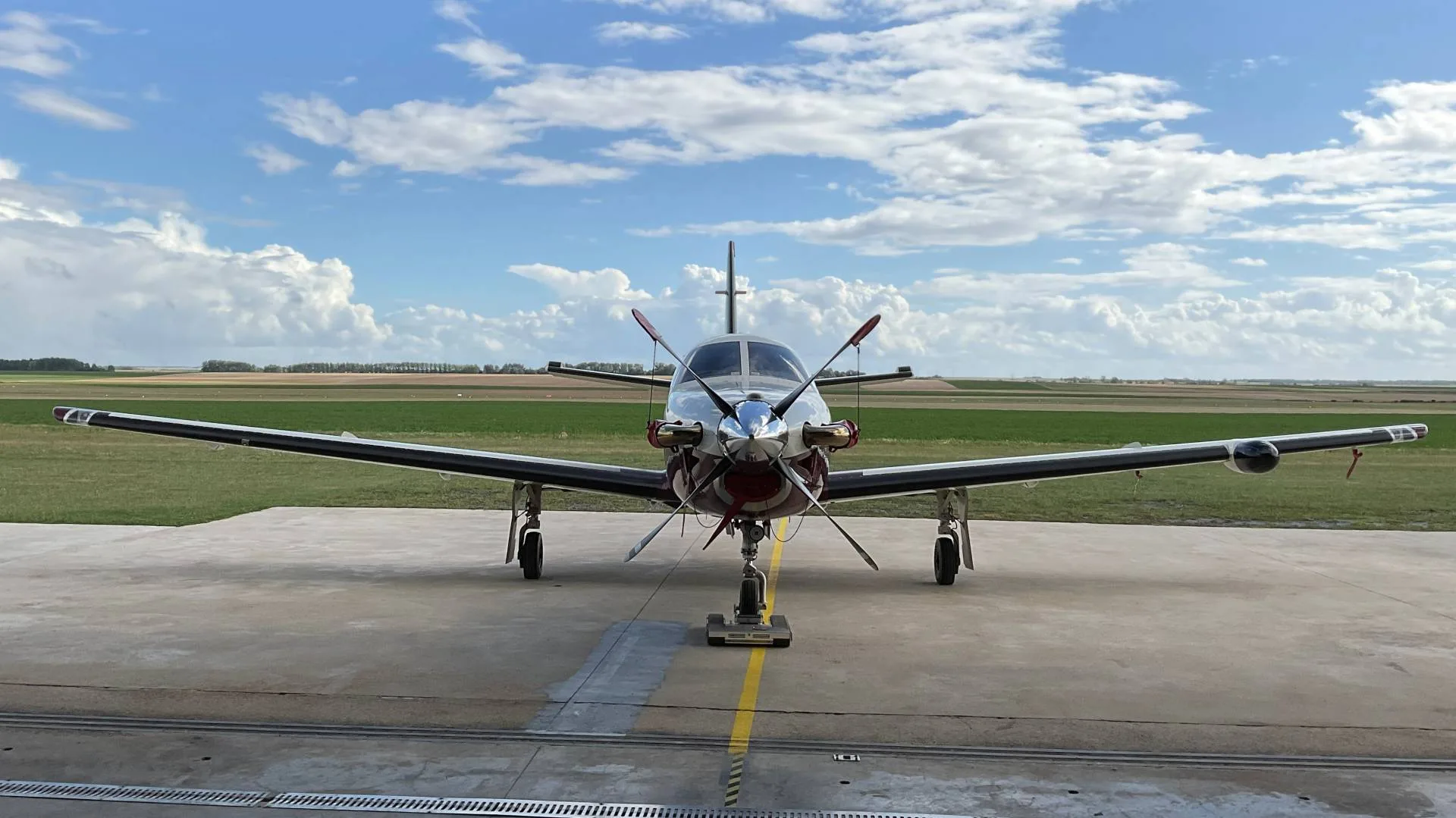 A silver and red Pilatus PC-12 turboprop aircraft is parked on a tarmac, viewed from the front, with fields and a cloudy sky in the background.