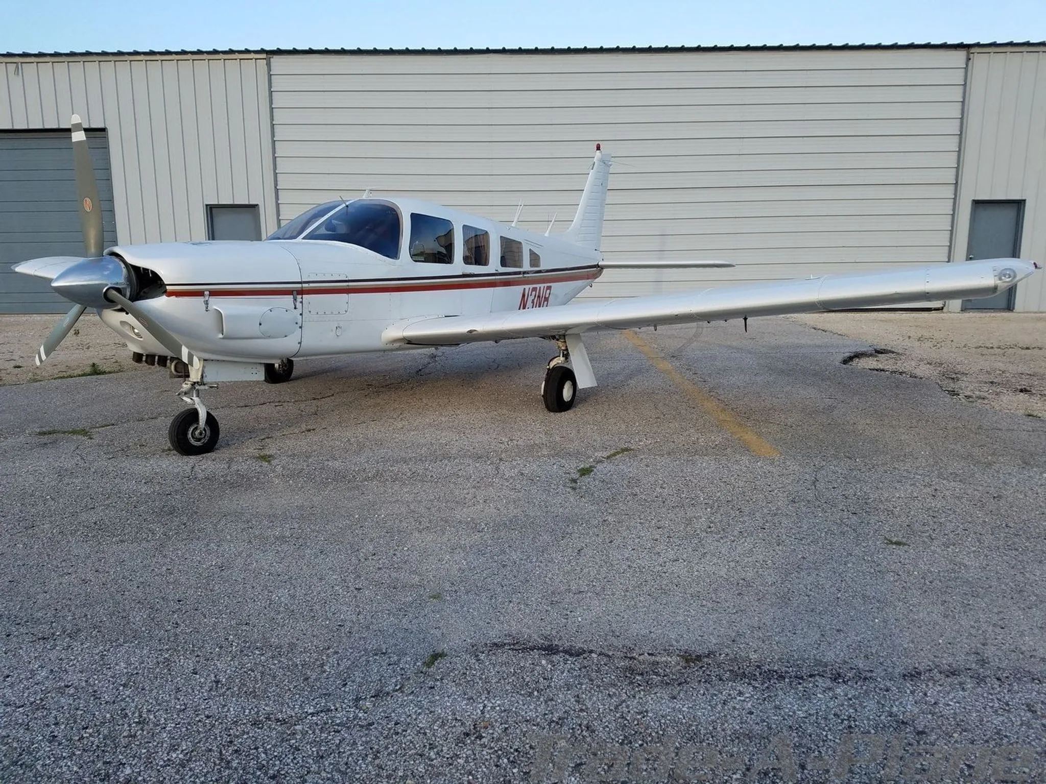 A white single-engine Piper aircraft with red and brown stripes, registration N3NB, parked in front of a metal hangar.