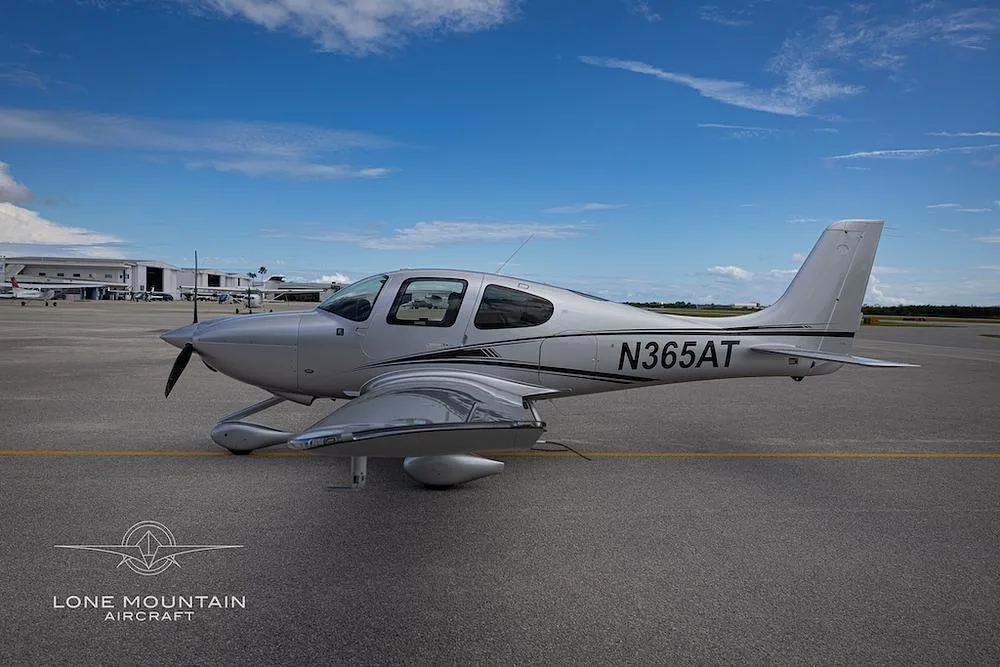 A silver Cirrus SR22 aircraft with black and white stripes, seen from the side on an airfield with hangars in the background.