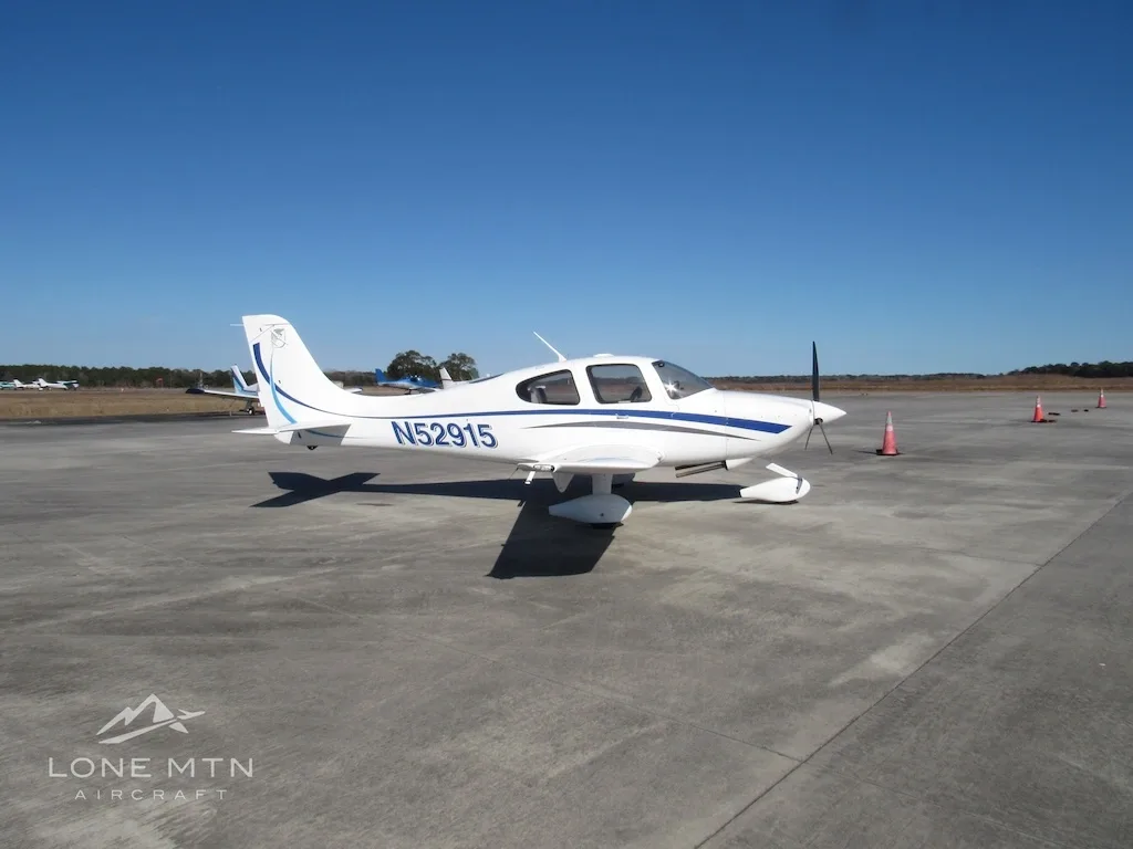 A white Cirrus SR20 aircraft with blue and grey stripes is parked on a concrete ramp, viewed from the front-left.