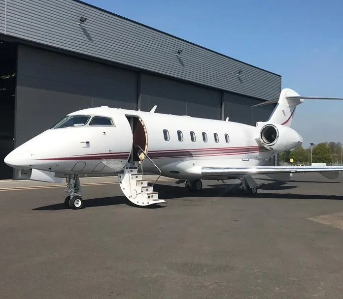 A white business jet with a maroon stripe, parked on an tarmac with its door open and stairs extended, next to a grey hangar.