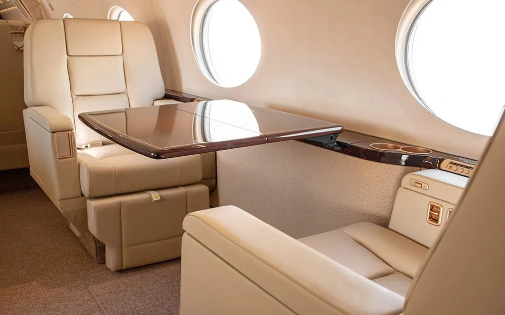 A luxurious aircraft interior with beige leather seats and a polished wooden table extended from the sidewall.