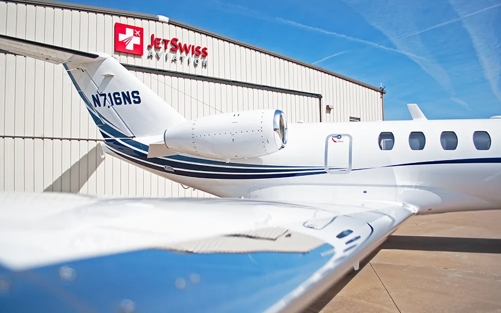 The tail section and wing of a white business jet with blue stripes and registration N716NS, parked outside a hangar.