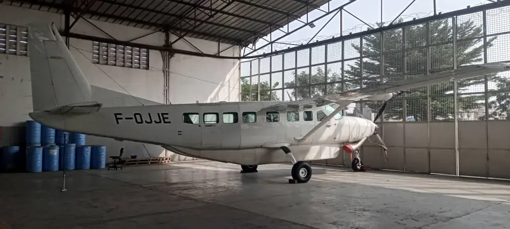 A white Cessna 208 Caravan aircraft with registration F-OJJE is parked inside a hangar, viewed from the rear left.