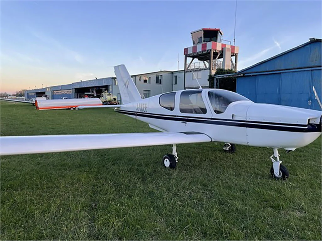 A white single-engine aircraft with black stripes parked on grass next to a runway, with a control tower and hangars in the background at sunset.