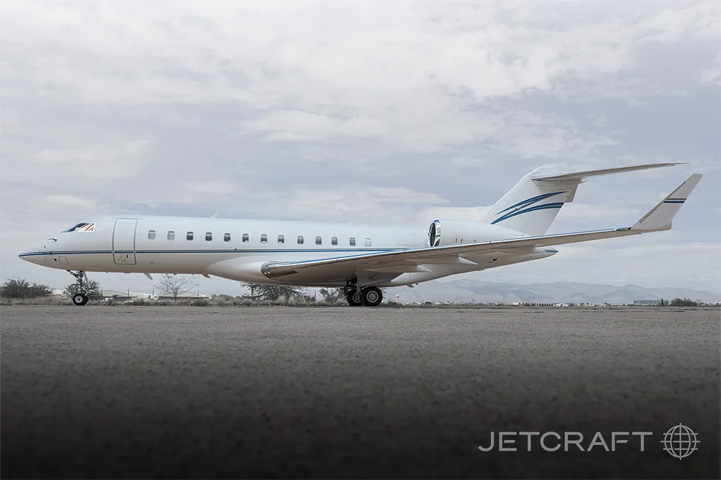 A white Bombardier Global 6000 business jet with blue and grey stripes on the tail and fuselage, parked on an airfield under a cloudy sky.