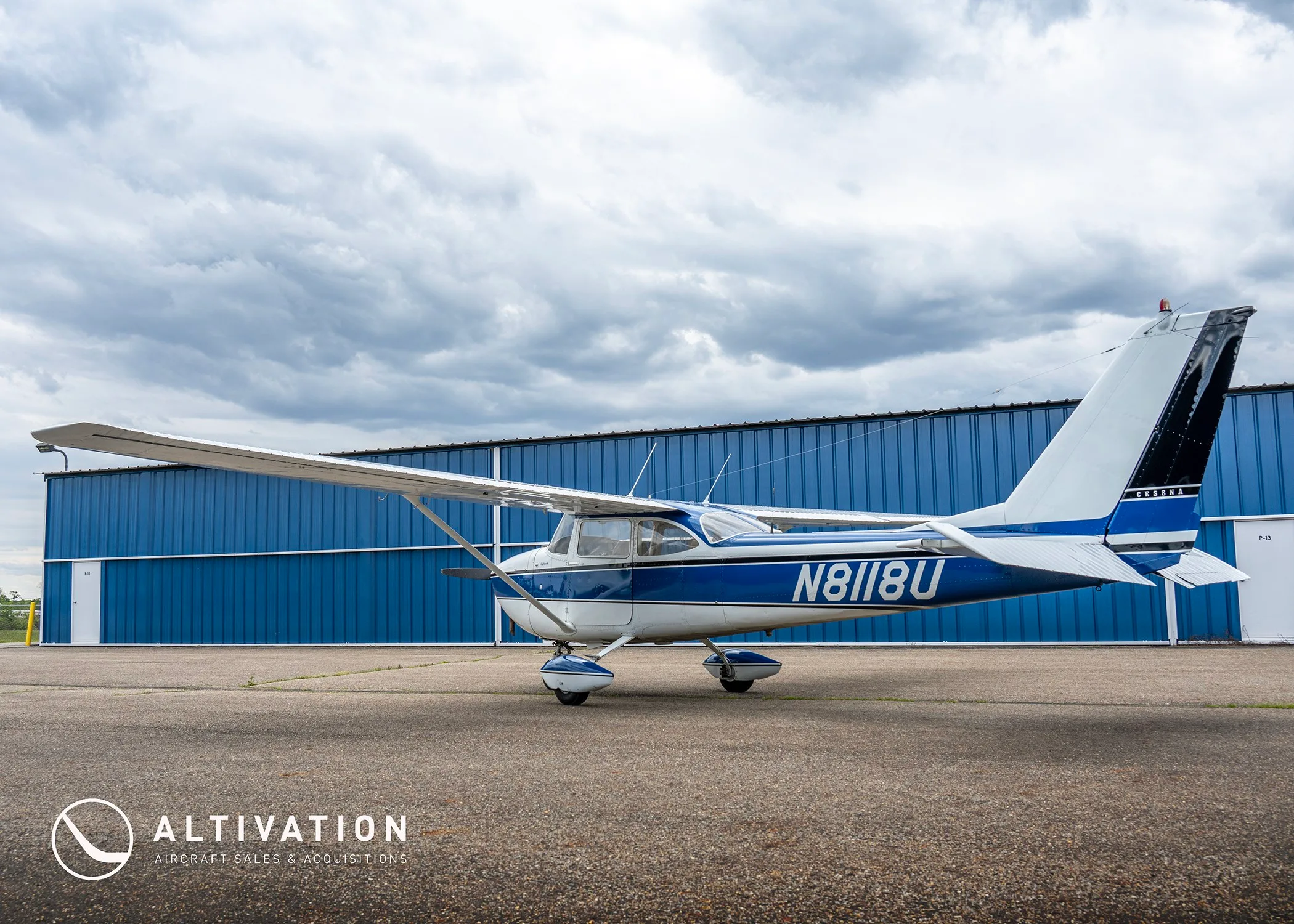 A rear-quarter view of the white and blue Cessna 172 Skyhawk N8118U parked on an asphalt ramp in front of a blue hangar.