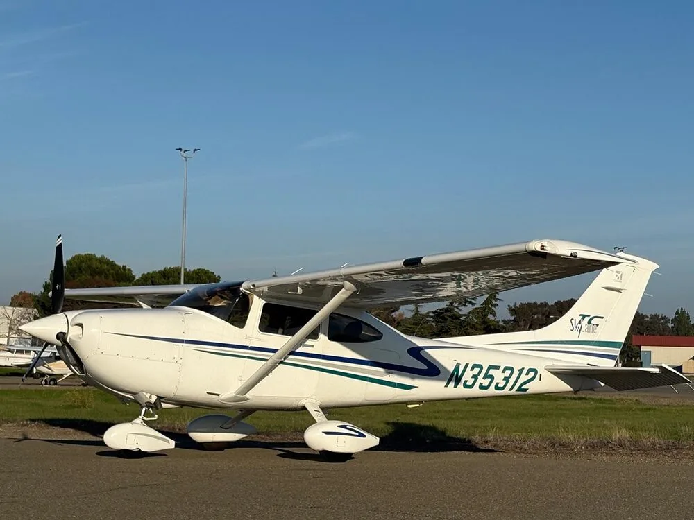 A white Cessna 182 Skylane with blue and green stripes, registration N35312, parked on an tarmac with trees and other aircraft in the background.