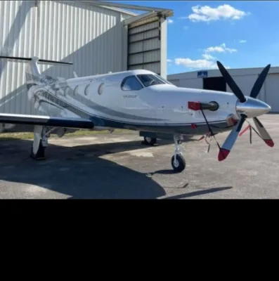 A white Pilatus PC-12 turboprop aircraft with grey stripes parked on an apron in front of a hangar.