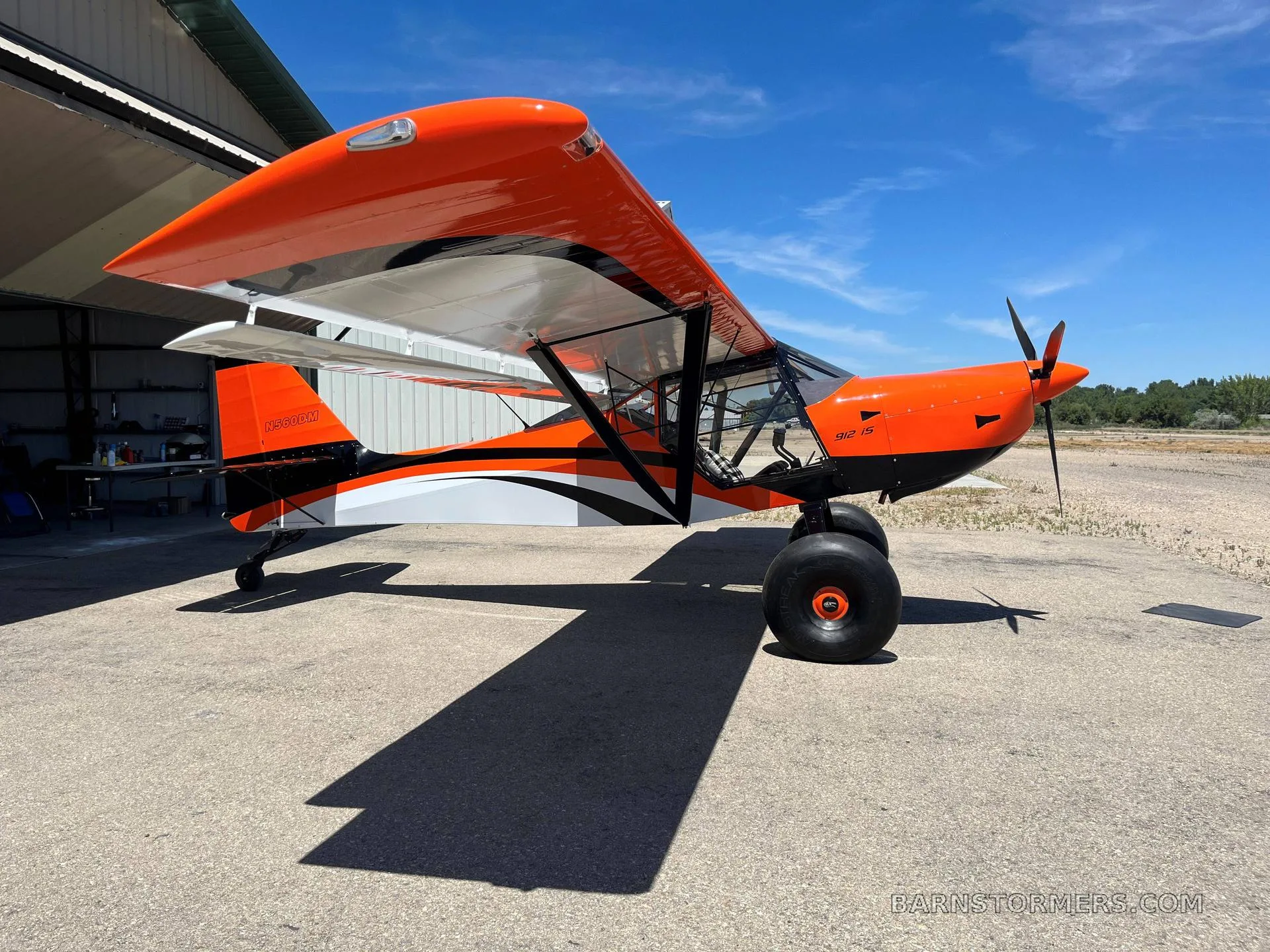 A bright orange and white Kitfox S7 Super Sport aircraft with large tires parked on a tarmac next to a hangar.