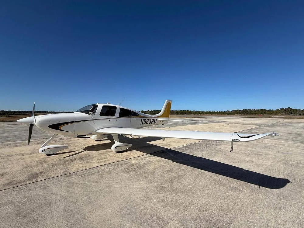A slightly angled side view of a white Cirrus SR22 aircraft with black and gold stripes and a gold tail, registration N583PU, on an airfield.