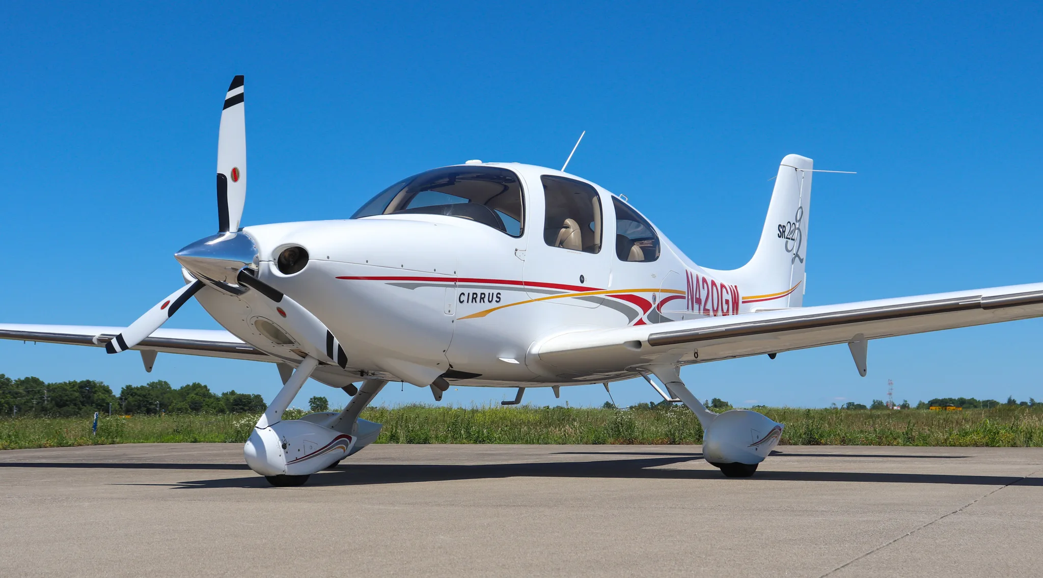 Front-left view of a white Cirrus SR22 aircraft with red and gold stripes on a tarmac under a clear blue sky.