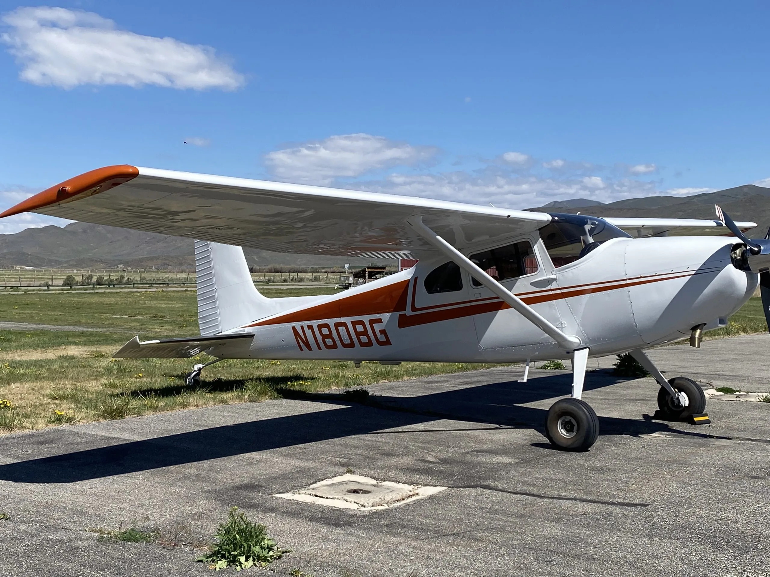 A white Cessna 170B with orange stripes and N180BG registration parked on an airfield with mountains in the background.