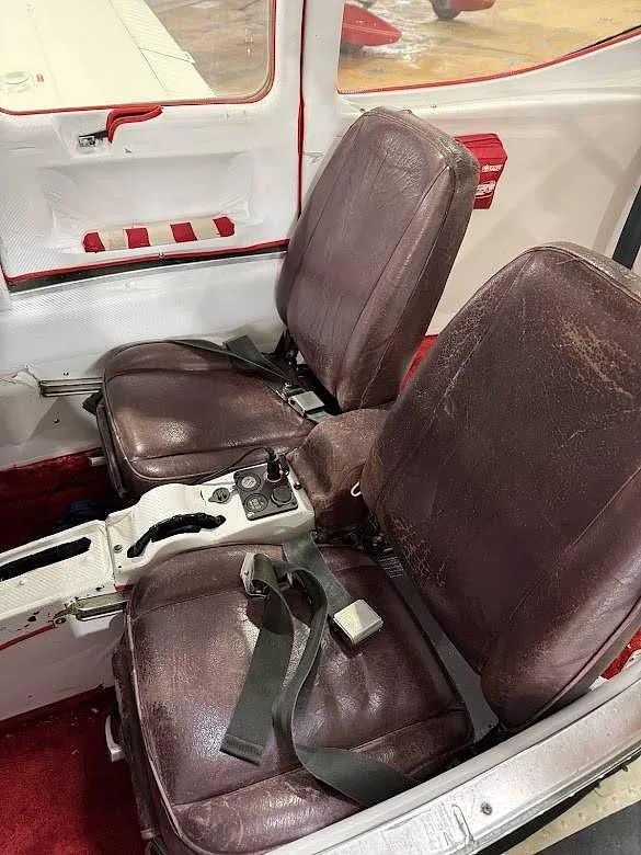 The interior cabin of a Piper Tomahawk aircraft, showing two brown leather seats and red carpet.