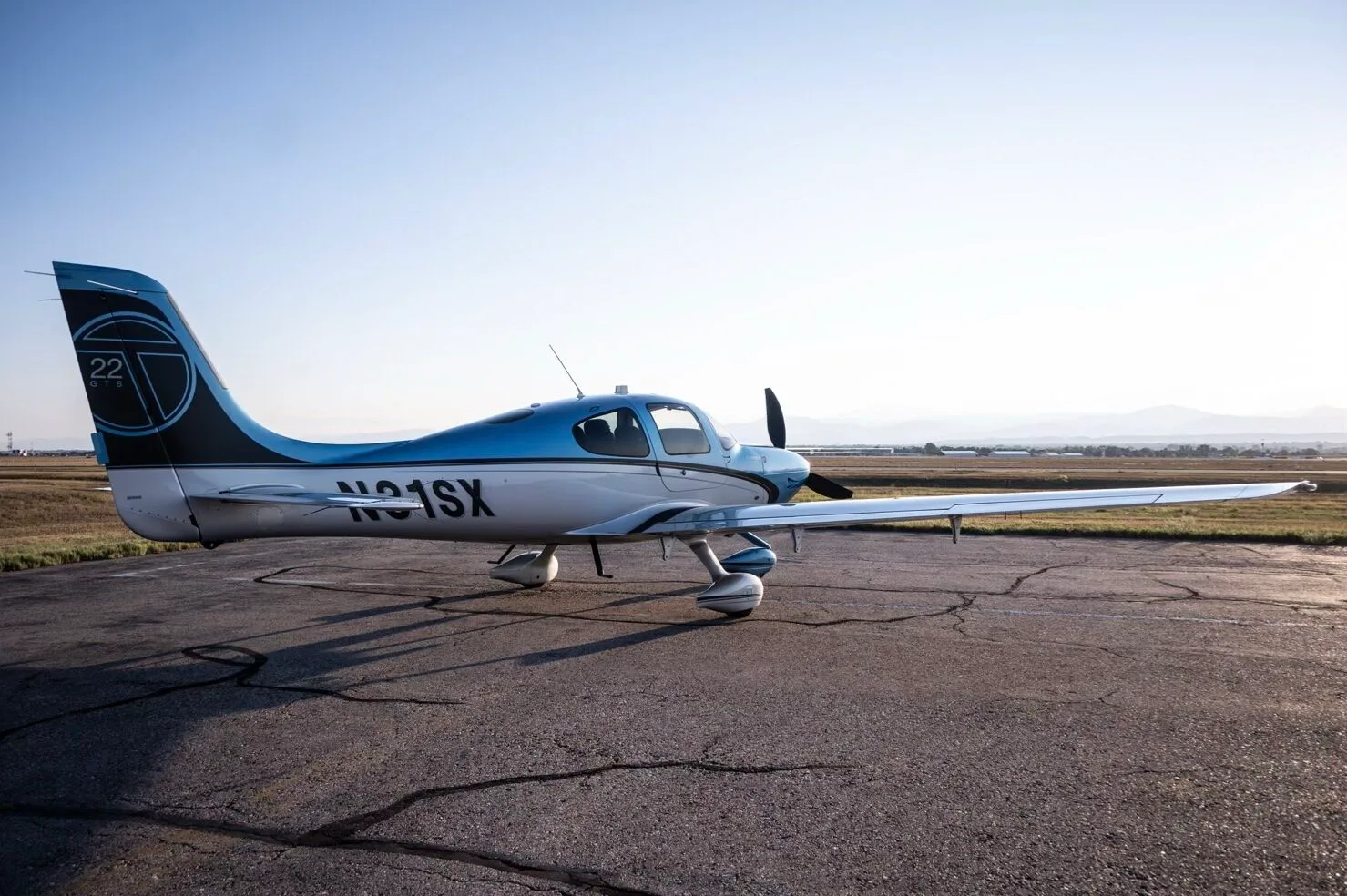 A Cirrus SR22T aircraft, N31SX, in blue and silver paint, parked on an airfield under a bright sky.