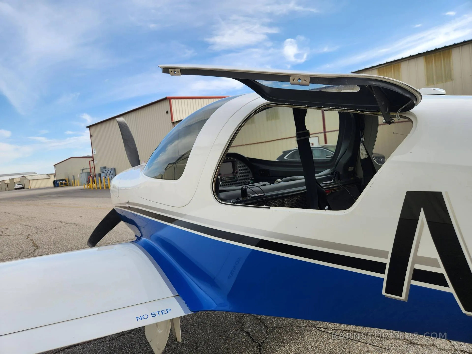 A side view of a white and blue aircraft with its canopy open, revealing the cockpit interior, parked on a tarmac.