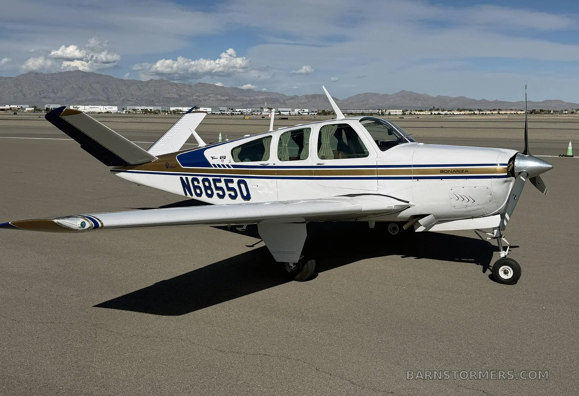 A white, blue, and gold Beechcraft Bonanza N68550 parked on an airfield with mountains in the background.