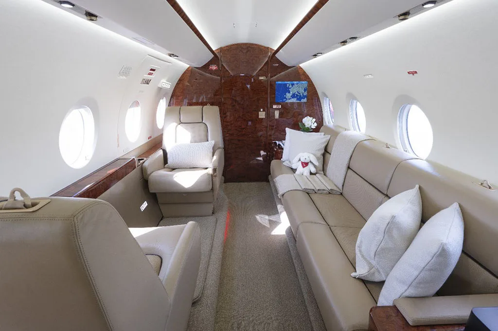 The luxurious interior cabin of a private jet, featuring beige leather seats, a couch, and polished wood accents.