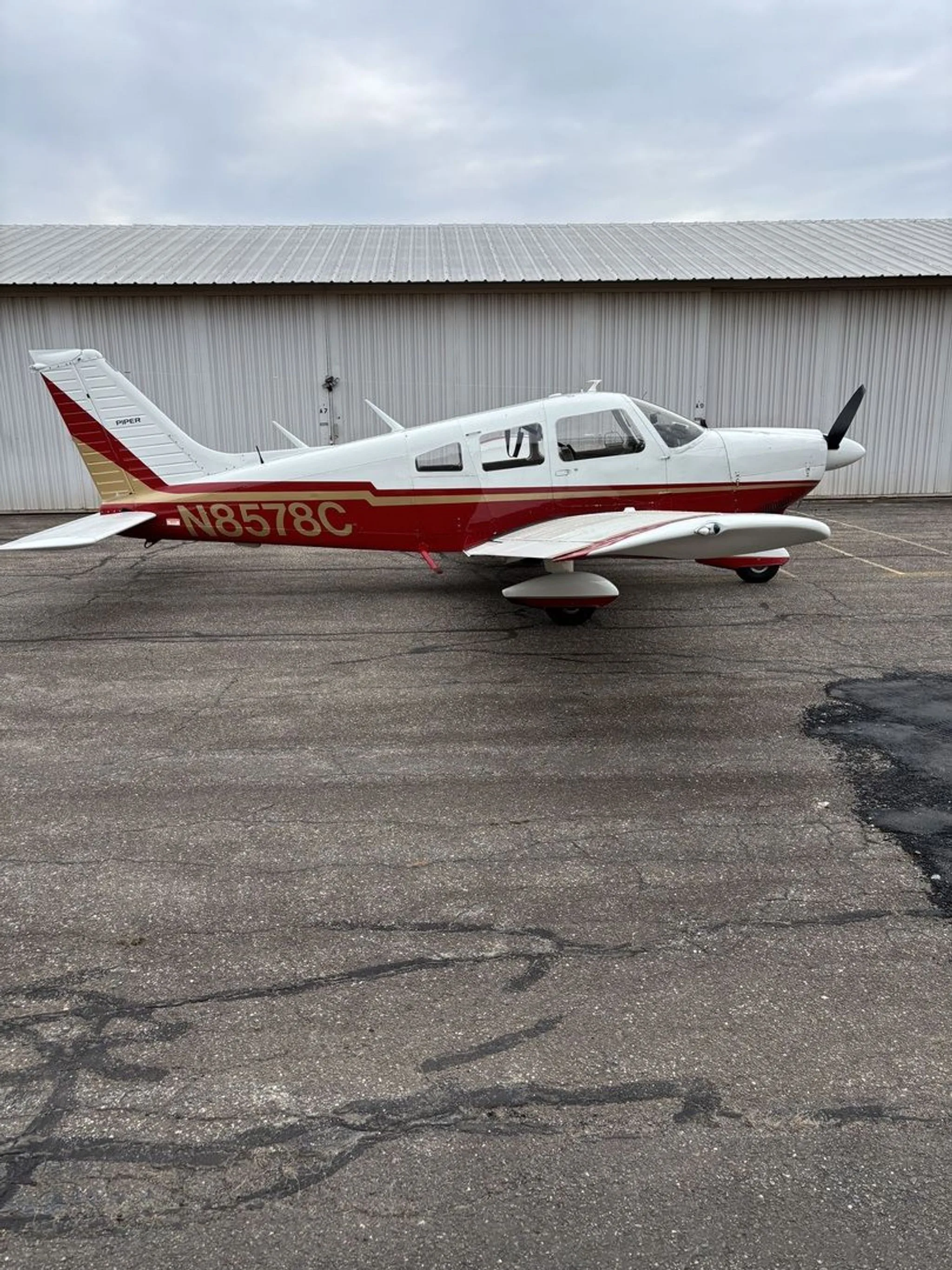A Piper aircraft with red and gold stripes over white paint, registration N8578C, parked on an asphalt apron next to a hangar.