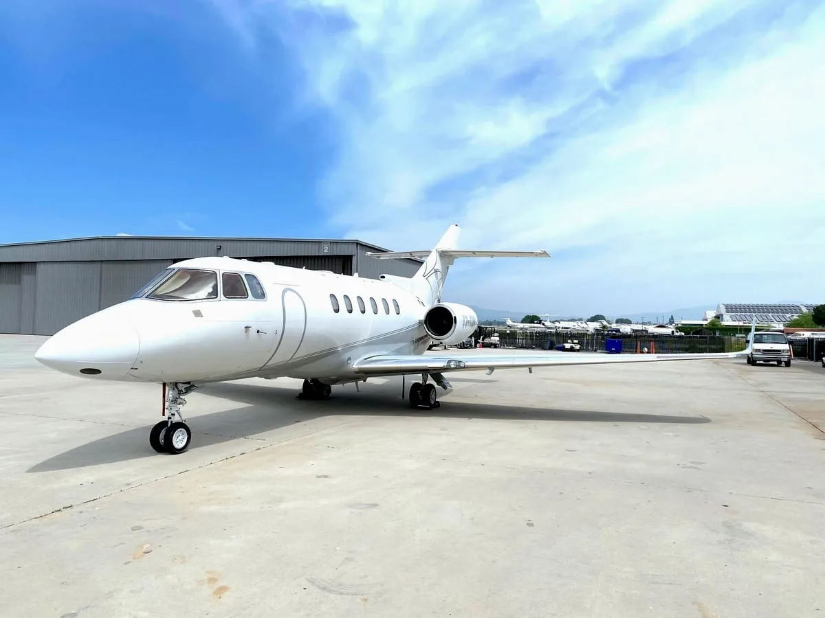 A white business jet, possibly a Hawker, parked on a tarmac next to a hangar under a blue sky with clouds.