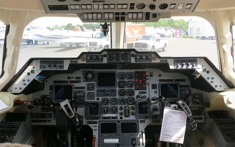 The cockpit of a business jet, showing the instrument panel, yokes, and overhead controls, with other aircraft visible outside.