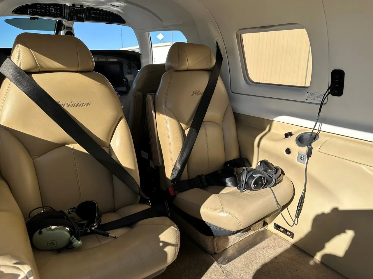 The tan leather passenger seats of a Piper Meridian aircraft cabin, with seatbelts fastened and headsets on the seats.
