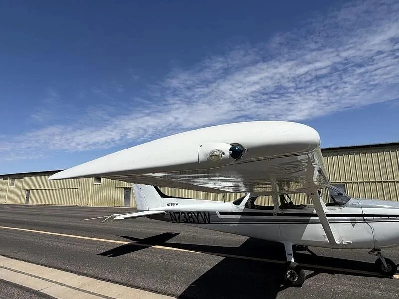 A side-right view of the white Cessna 172, N738YW, highlighting the wing and fuselage, parked on a tarmac.