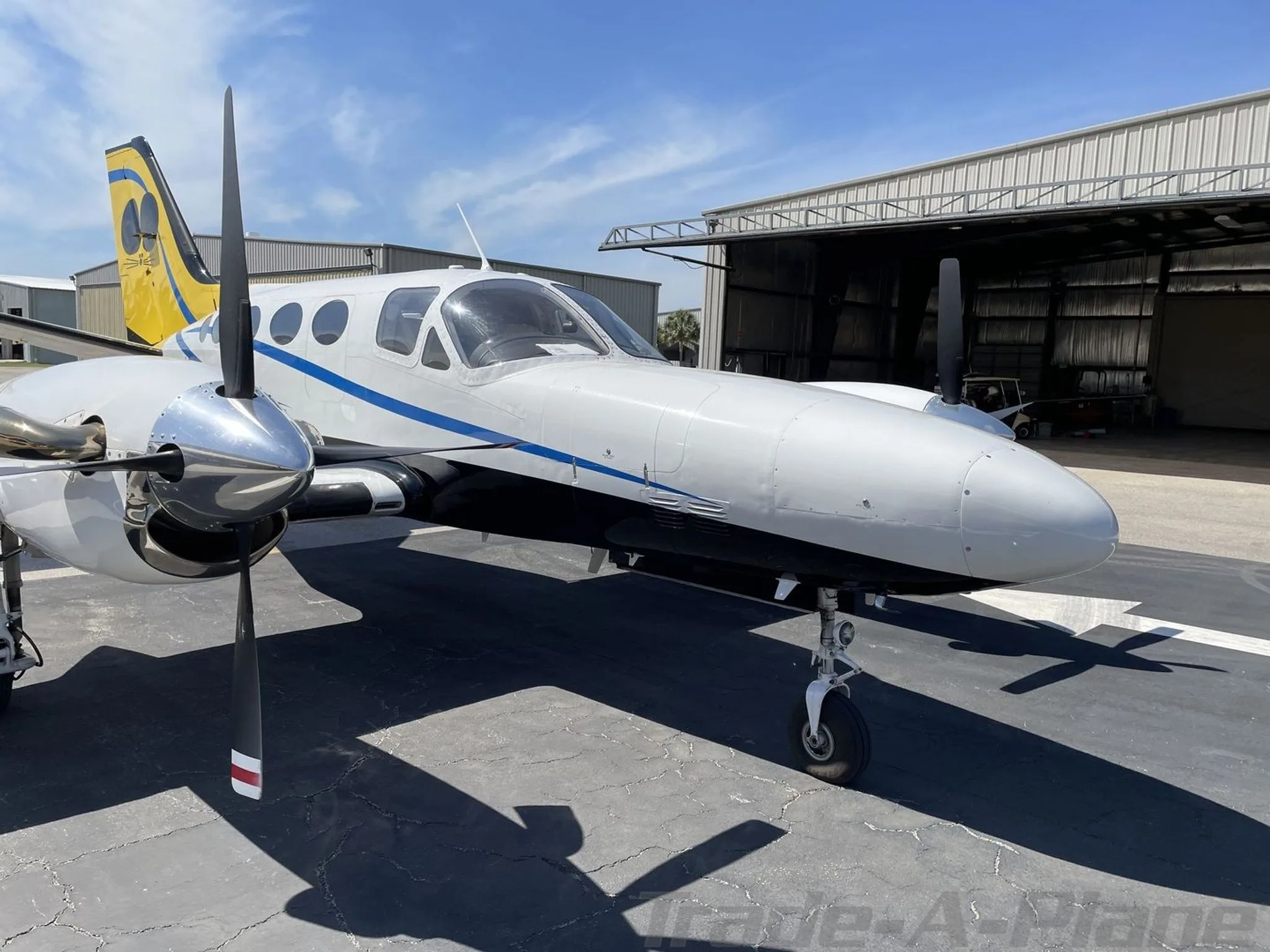 A front-right view of a white and blue Cessna 441 Conquest II with a yellow and black tail, parked on a tarmac next to a hangar.