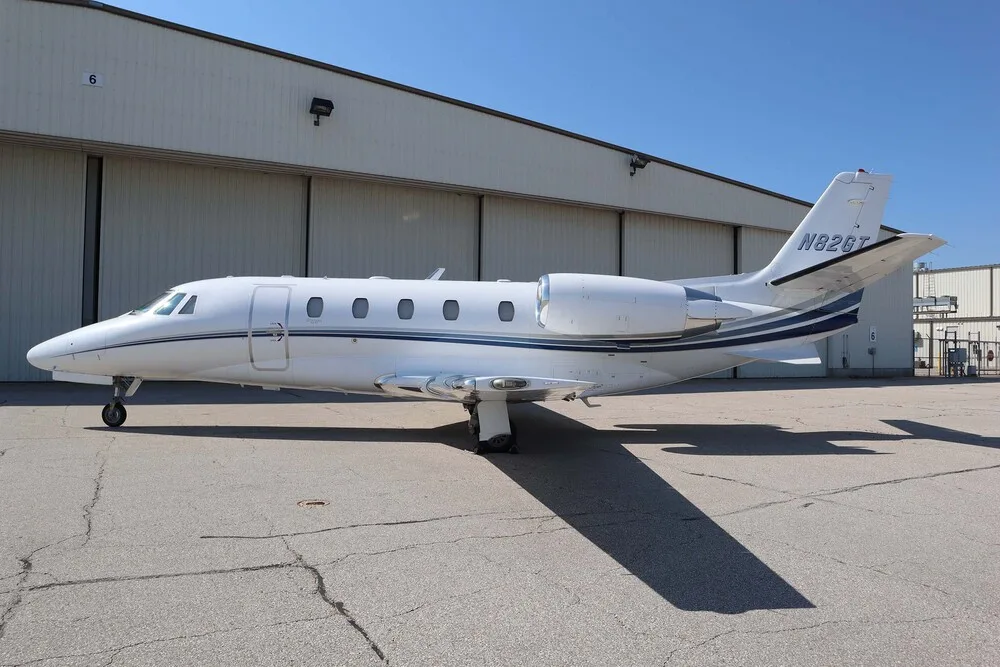 A white Cessna Citation business jet with blue and grey stripes, registration N82GT, parked on a tarmac next to a hangar.
