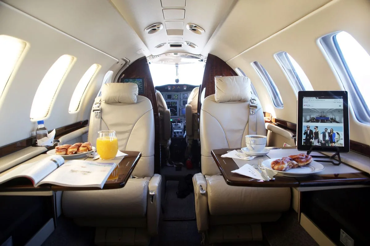 The luxurious interior cabin of a private jet, featuring beige leather seats, fold-out tables with breakfast items, and a tablet displaying a group of people.