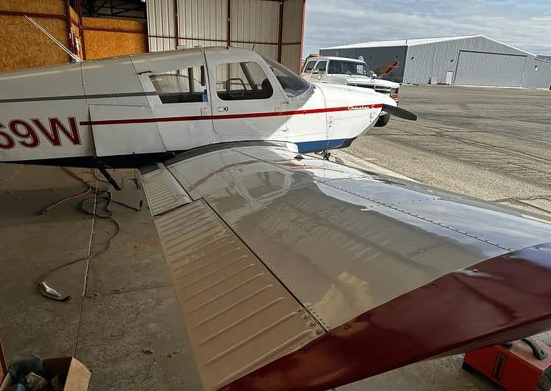 A close-up view of the wing and fuselage of a white Piper Cherokee C aircraft, partially in a hangar with other buildings in the background.